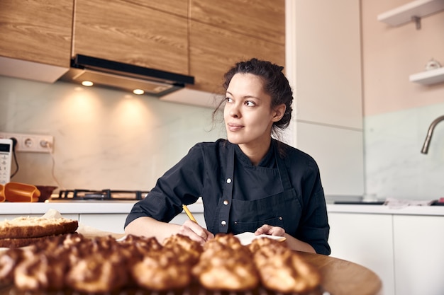 Cozinheira profissional está escrevendo uma receita de éclairs na moderna cozinha clara