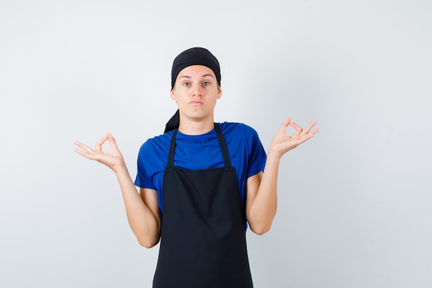 Cozinheiro adolescente masculino mostrando gesto de meditação em t-shirt, avental e olhando confiante, vista frontal.