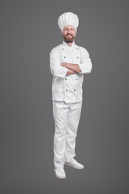 Cozinheiro alegre vestindo uniforme branco e boné em pé com os braços cruzados em um fundo cinza no estúdio