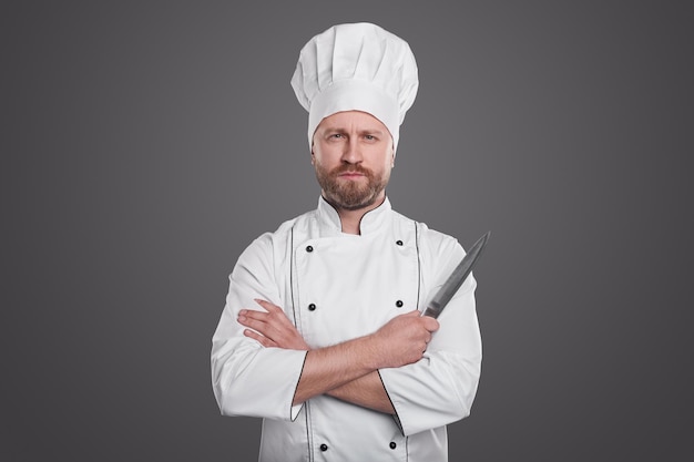 Cozinheiro barbudo confiante em um uniforme branco em pé com uma faca em um fundo cinza e olhando para a câmera