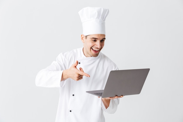 Cozinheiro chef alegre vestindo uniforme em pé sobre uma parede branca, segurando um laptop