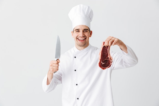 Cozinheiro chef alegre vestindo uniforme mostrando bife cru segurando uma faca isolada na parede branca