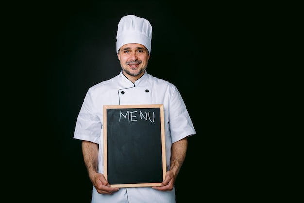 Cozinheiro chefe de jaqueta e chapéu de cozinha sorrindo, segurando uma piazarra com o menu, sobre fundo preto