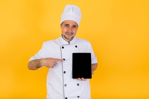 Cozinheiro chefe vestindo jaqueta de cozinha branca e chapéu, segurando um tablet e apontando um dedo, sobre fundo amarelo