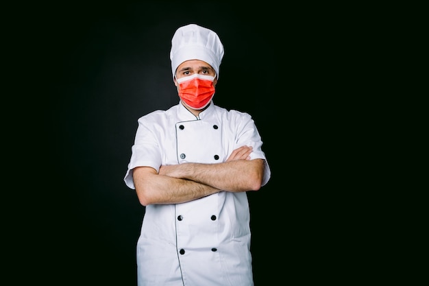 Cozinheiro chefe vestindo jaqueta e chapéu, com uma máscara vermelha, braços cruzados, sobre fundo preto