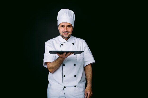 Cozinheiro chefe vestindo jaqueta e chapéu, segurando uma placa de ardósia, sobre fundo preto
