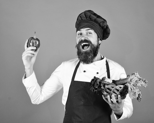 Cozinheiro com rosto alegre em uniforme cor de vinho segura pimenta e legumes em tigela de vime Conceito de mercearia verde Homem com barba em fundo azul Chef segura alface, tomate, pimentão e cogumelos