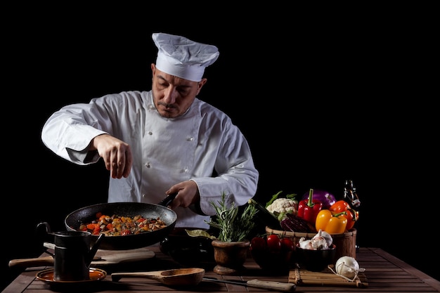 Cozinheiro de uniforme branco e chapéu colocando sal e ervas no prato de comida com legumes antes de servir enquanto trabalhava na cozinha de um restaurante