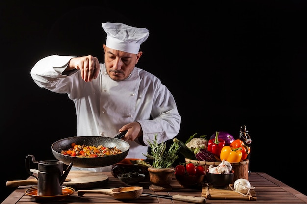 Cozinheiro masculino de uniforme branco e chapéu colocando sal e ervas no prato de comida com vegetais