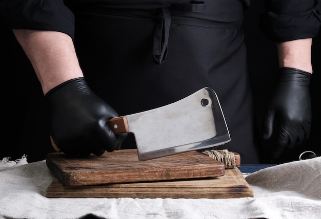 Cozinheiro masculino em uniforme preto e luvas de látex pretas mantém uma faca de carne afiada grande sobre uma tábua