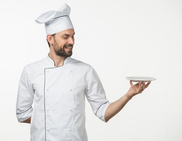 Cozinheiro masculino profissional no uniforme branco do cozinheiro chefe que apresenta o prato no fundo branco