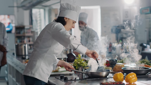 Cozinheiro masculino usando frigideira para cozinhar legumes a vapor para prato gourmet, cozinhando comida deliciosa com fumaça na cozinha profissional do restaurante. Homem de uniforme branco, preparando a refeição cozida. Tiro de mão.