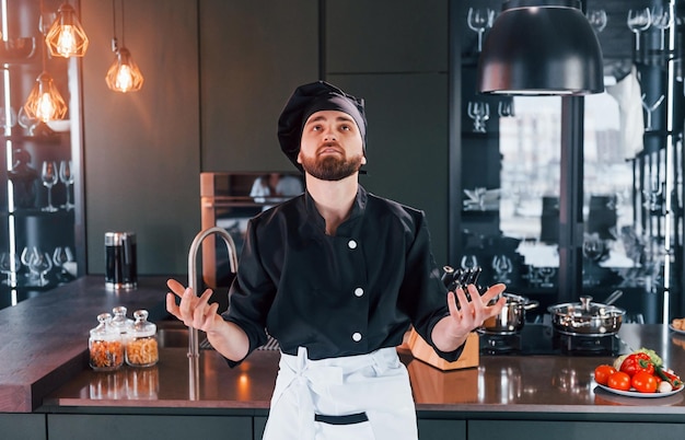 Cozinheiro profissional jovem chef de uniforme em pé perto da mesa e posando para uma câmera na cozinha