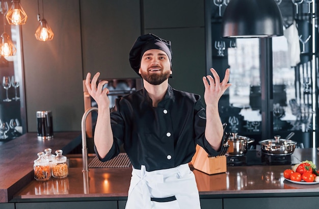 Cozinheiro profissional jovem chef de uniforme em pé perto da mesa e posando para uma câmera na cozinha