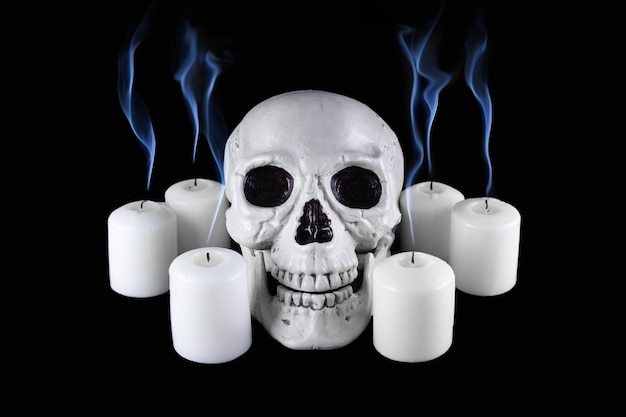 Cráneo humano entre velas blancas apagadas con columnas de humo azul en la oscuridad, naturaleza muerta, altar.