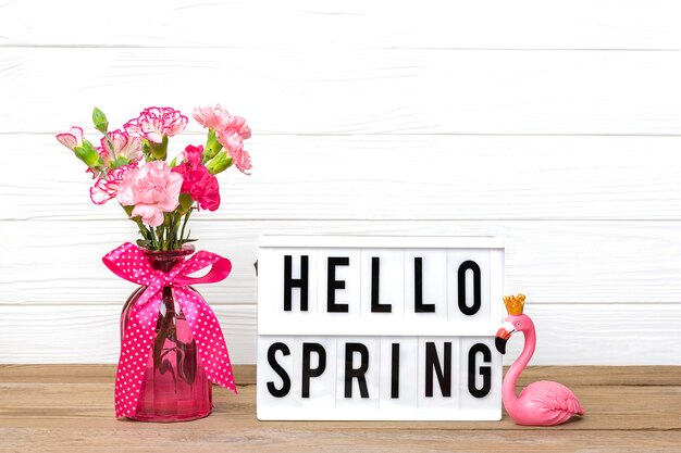 Cravos cor de rosa pequenos em um vaso e caixa de luz com texto olá primavera, figura de flamingo na