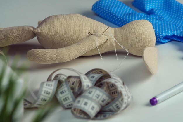 Creación de costura de juguete de conejo de peluche con aguja en el cuerpo acostado sobre la mesa Juguete de liebre de hobby de punto cerca de tejido de lunares azules