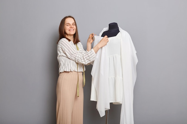 Creaciones de ropa personalizada Emprendedor de moda innovador Artesanía en diseño textil Mujer positiva costurera de pie cerca de un vestido nuevo en maniquí aislado sobre fondo gris