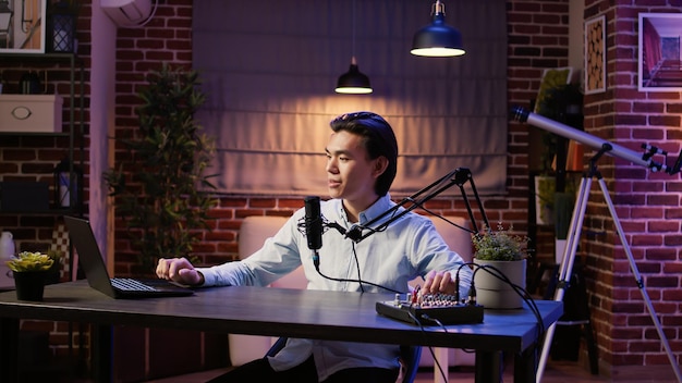 Creador de contenido asiático hablando con la audiencia en un episodio de podcast en línea, grabando conversaciones para los oyentes del canal. Uso de equipos de podcast para redes sociales, transmisión de audio en vivo.