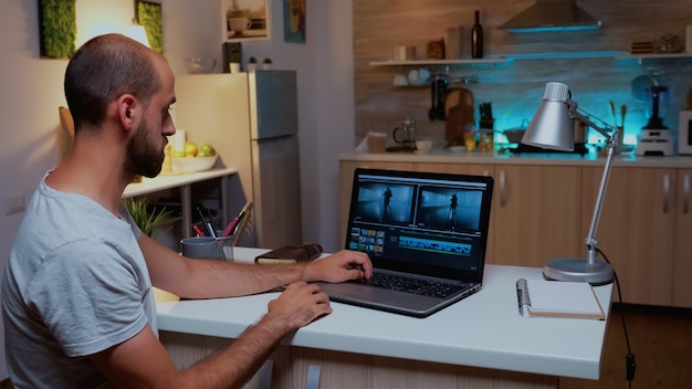 Foto creador de contenido que trabaja de forma remota en una computadora portátil desde casa durante la noche. videógrafo de montaje de película de audio de edición en portátil profesional sentado en un escritorio en la cocina moderna a medianoche