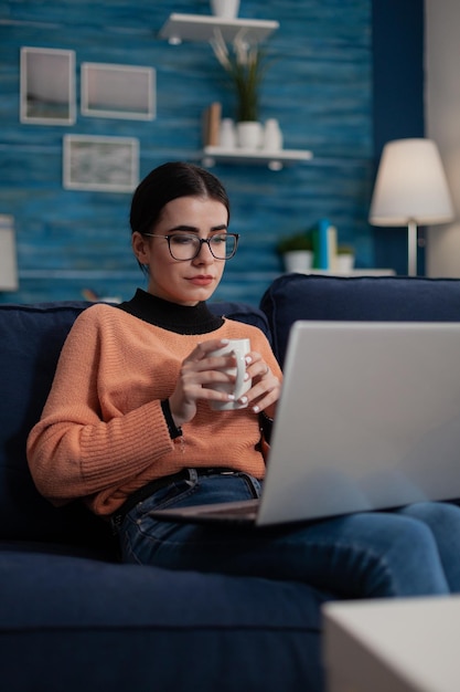 Creador de contenido en el sofá con una computadora portátil en el regazo sosteniendo una taza. Estudiante con anteojos sentado en un sofá aprendiendo a distancia desde casa en la sala de estar. Influencer relajándose mirando la pantalla.