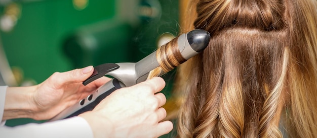 Creando rizos con tenacillas. Peluquería hace un peinado para una mujer joven con largo cabello rojo en un salón de belleza.