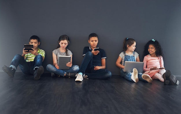 Creciendo en la era digital Foto de estudio de niños sentados en el suelo y usando tecnología inalámbrica contra un fondo gris