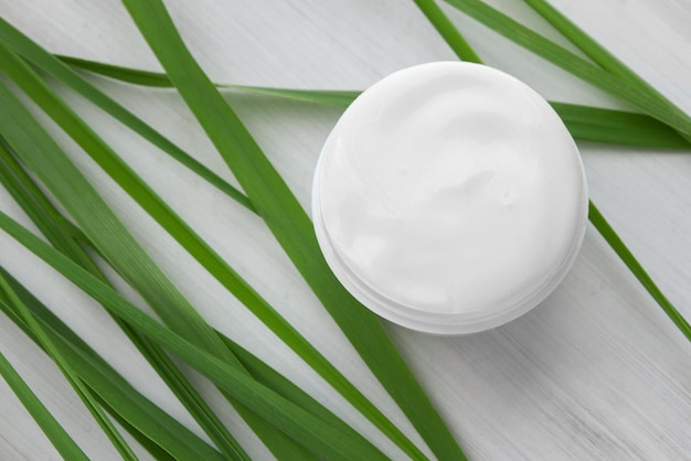 Crema cosmética blanca sobre un fondo de madera blanca rodeada de largas hojas verdes