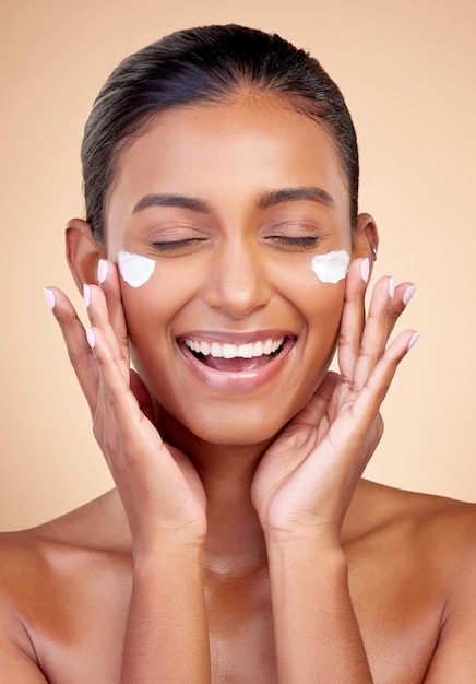 Crema facial y mujer feliz para belleza cosméticos dermatología o maquillaje estético en fondo de estudio modelo femenina india loción y sonrisa con protector solar facial para cuidado de la piel limpia brillo y brillo