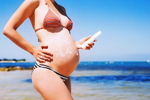 Crema hidratante para el cuidado de la piel de la mujer embarazada en el vientre en la playa del mar en verano Primer plano de un hermoso vientre embarazado