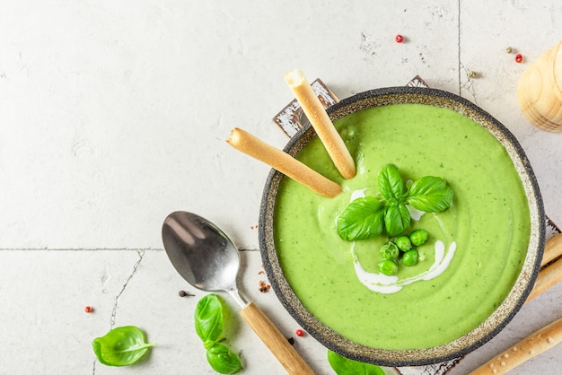 Crema verde saludable de guisantes y brócoli con albahaca y palitos de pan italiano crujiente grissini en un recipiente con una cuchara sobre la superficie de piedra clara