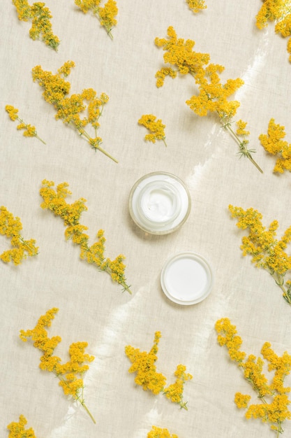 Creme cosmético em recipiente redondo e flores silvestres de ervas amarelas em têxteis naturais