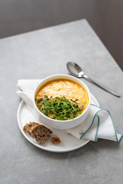 Creme de sopa de cenoura em uma tigela branca Tigela de sopa de cenoura com folhas de rúcula na mesa de concreto