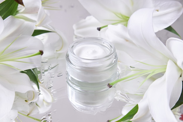 Creme zur Hautpflege, Naturkosmetik aus Blumen und Blütenblättern. Zwischen den Lilienblüten steht ein Glas mit weißer Creme