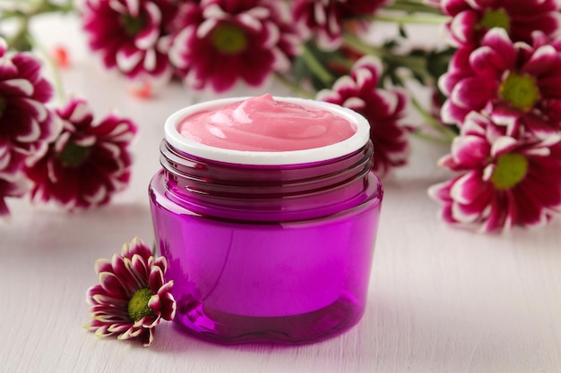 Cremekosmetik für Gesicht und Körper Rosafarbene Creme und Blume in einem violetten Glas auf einem weißen Holztisch