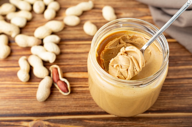 Cremige Erdnusspaste im offenen Glas, Erdnussbutter im Löffel. Erdnüsse in der Schale verstreut auf dem braunen Holztisch.