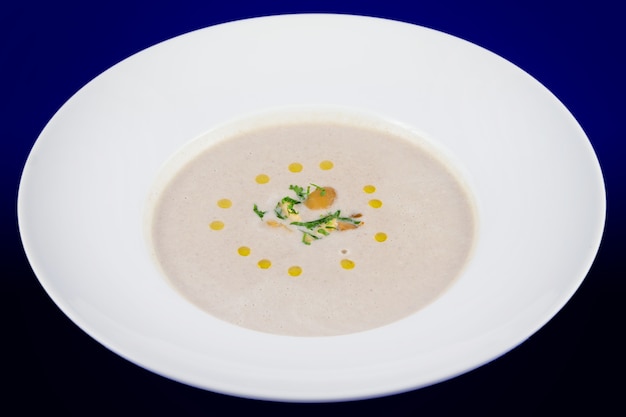 Cremige Pilzsuppe mit Kräutern auf einem weißen Teller und einem blauen Tisch.