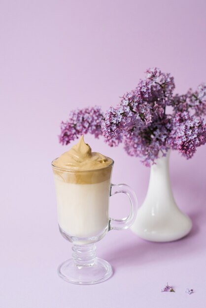 Cremiger Kaffee in Glastasse und lila Blüten in weißer Vase auf hellviolettem Hintergrund