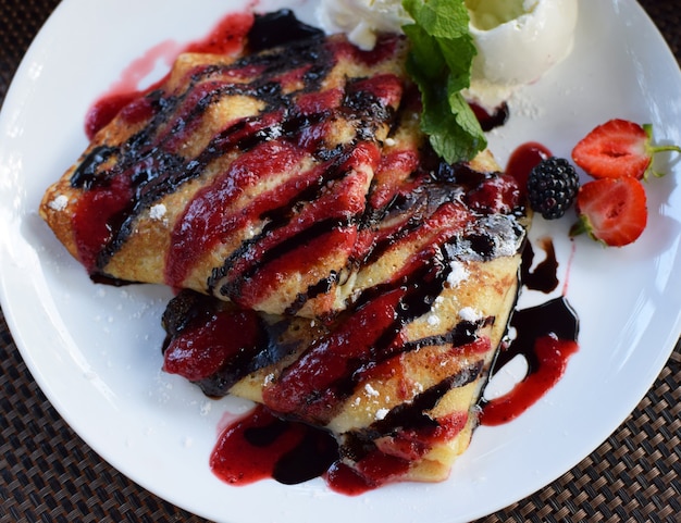 Crepas rellenas de fruta y cubiertas con siropes de chocolate y frutos rojos. Servido con bolas de helado y fresas. Tortitas finas, blini. Vista superior