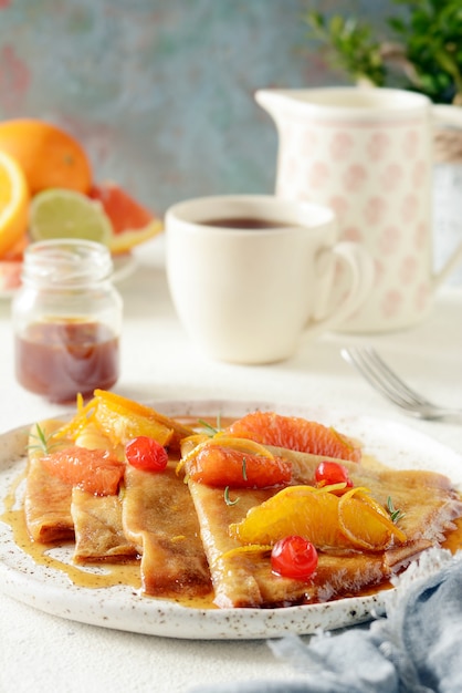 Crêpe Suzette zum Frühstück mit Orangen-Karamell-Sauce, Orangenscheiben, Limetten- und Orangenschale und einer Tasse Kaffee