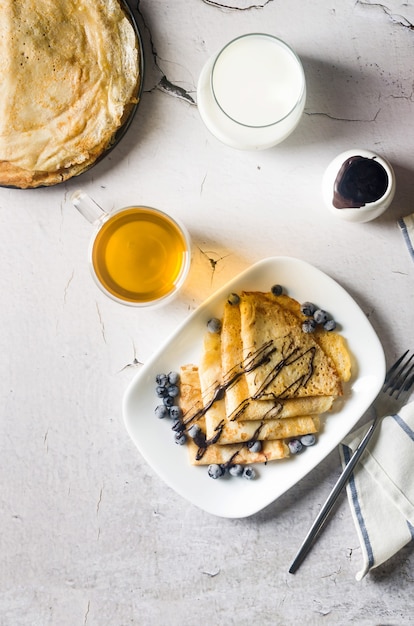 Crepes caseros, deliciosos panqueques finos con chocolate y bayas en un plato blanco para desayunar con una taza de té. Cocina tradicional rusa.