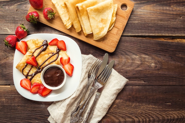 Crepes caseros frescos con fresas y salsa de chocolate sobre fondo de madera rústica. Vista superior.