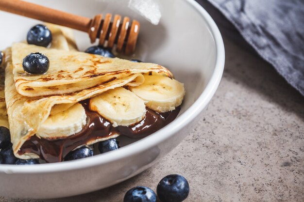 Crepes con chocolate, plátano, bayas y miel en un plato blanco.