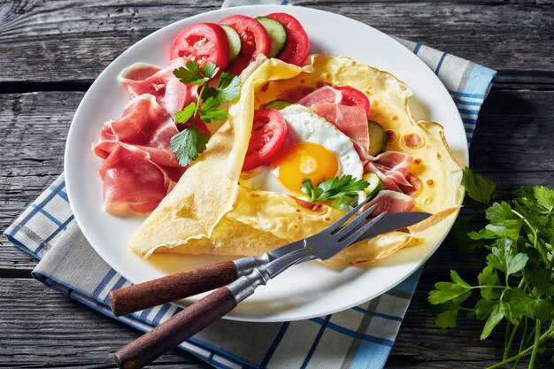 Crepes com ovo frito, fatias de Jamon, tomates frescos e recheios de pepino em um prato branco sobre uma velha mesa de madeira, vista de cima