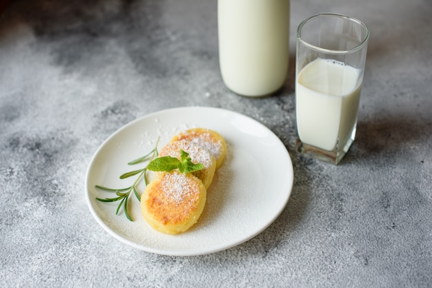 Crepes frescas sabrosas del requesón en una placa blanca con un vidrio de leche en un fondo concreto. Desayuno saludable y dietetico.