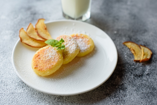 Crepes frescas sabrosas del requesón en una placa blanca con un vidrio de leche en un fondo concreto. Desayuno saludable y dietetico.