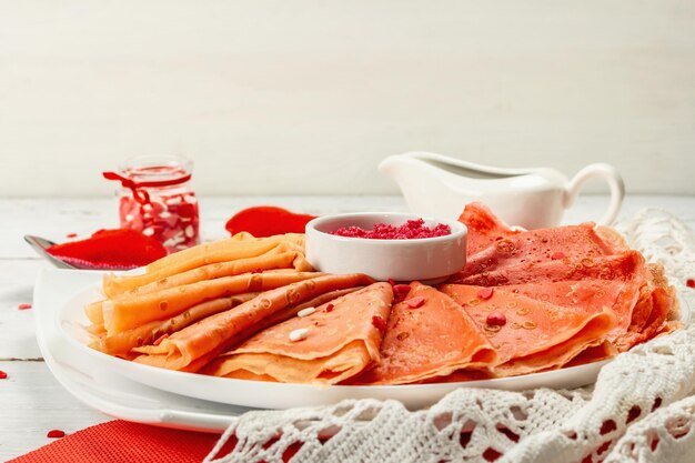 Crepes ou panquecas vermelhas finas com açúcar de framboesa e cobertura de pastelaria de corações de doce. Café da manhã romântico para o dia dos namorados ou das mães. Comida de gradiente de cor vermelha, fundo branco de madeira, close-up