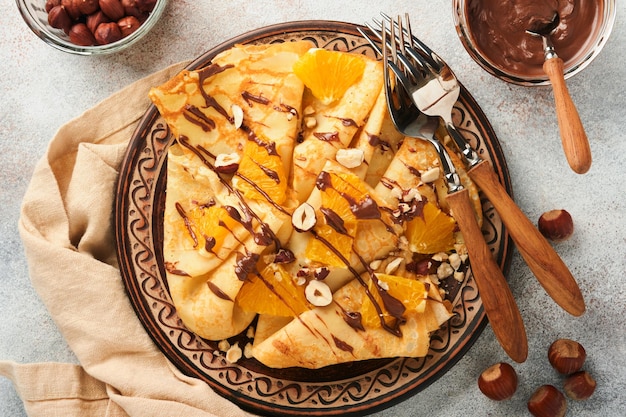 Crepes Suzette mit Orangen und Schokolade Dünne Crêpes mit Schokoladenaufstrich, Haselnüssen und Orangenscheiben Obst im Teller zum Frühstück auf altem rustikalem Tischhintergrund aus Beton Kopierbereich Ansicht von oben