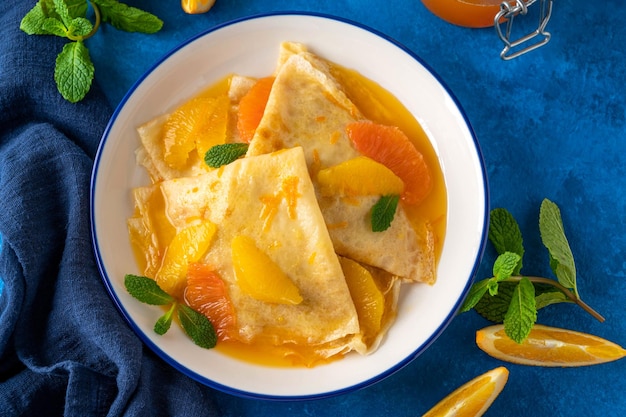 Crêpes-Suzette mit Orangensirup und Scheibenfrüchten auf einem Teller auf einer blauen Betonhintergrund-Draufsicht