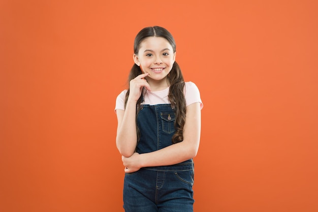 Crezca un niño mental y físicamente sano. Bienestar y salud. Concepto de infancia. Chica de moda. Niño adorable niña de pie sobre fondo naranja. Cuál es la clave de la felicidad infantil. Infancia feliz.